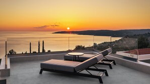 Une terrasse sur le toit offrant une vue sur le coucher de soleil sur la mer, une balustrade en verre et un espace salon moderne.