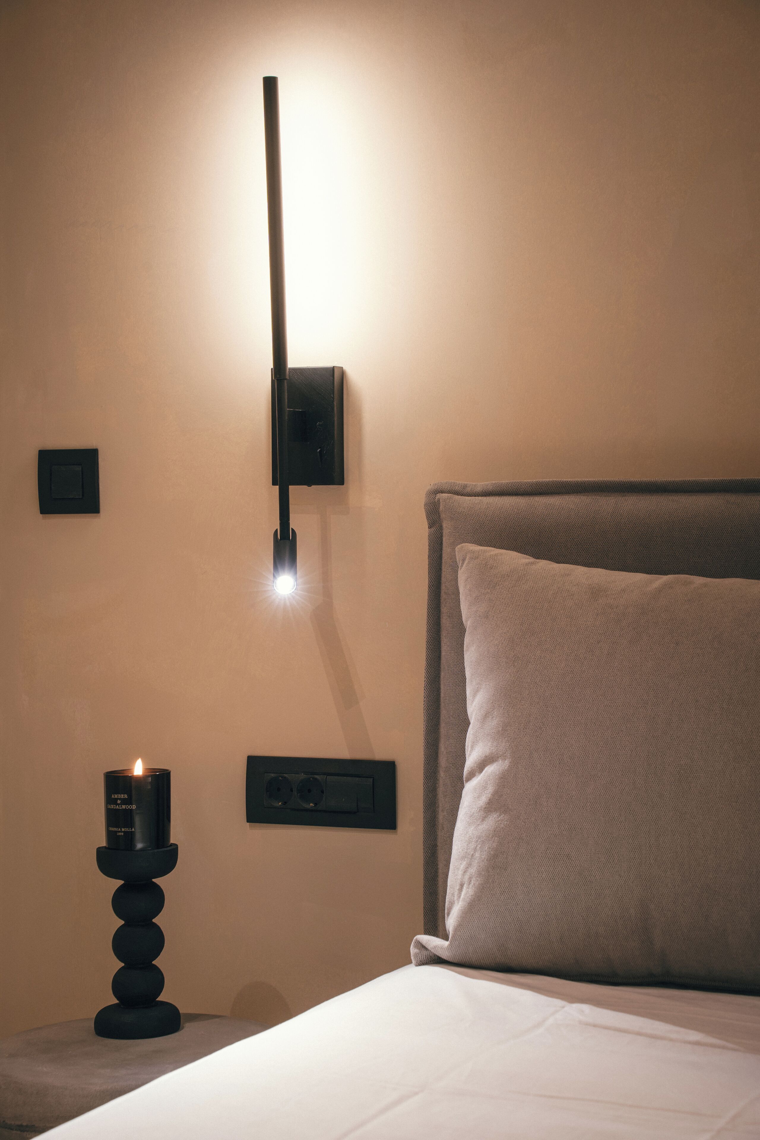A neatly made bed with a headboard, a wall-mounted light fixture, and a candle on a stand.