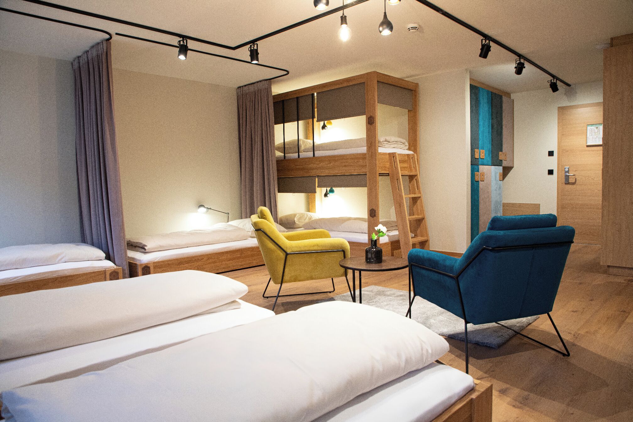 A hotel room with bunk beds, a yellow armchair, a blue armchair, a small table with a vase, and a ladder.