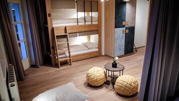 Una habitación con literas, una mesita con una planta, dos taburetes de mimbre y un radiador.