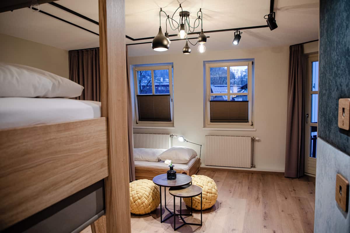 Ein Zimmer mit einem Etagenbett, einem Einzelbett, einem Tisch mit Lampe, zwei gewebten Poufs und zwei Fenstern mit Jalousien.