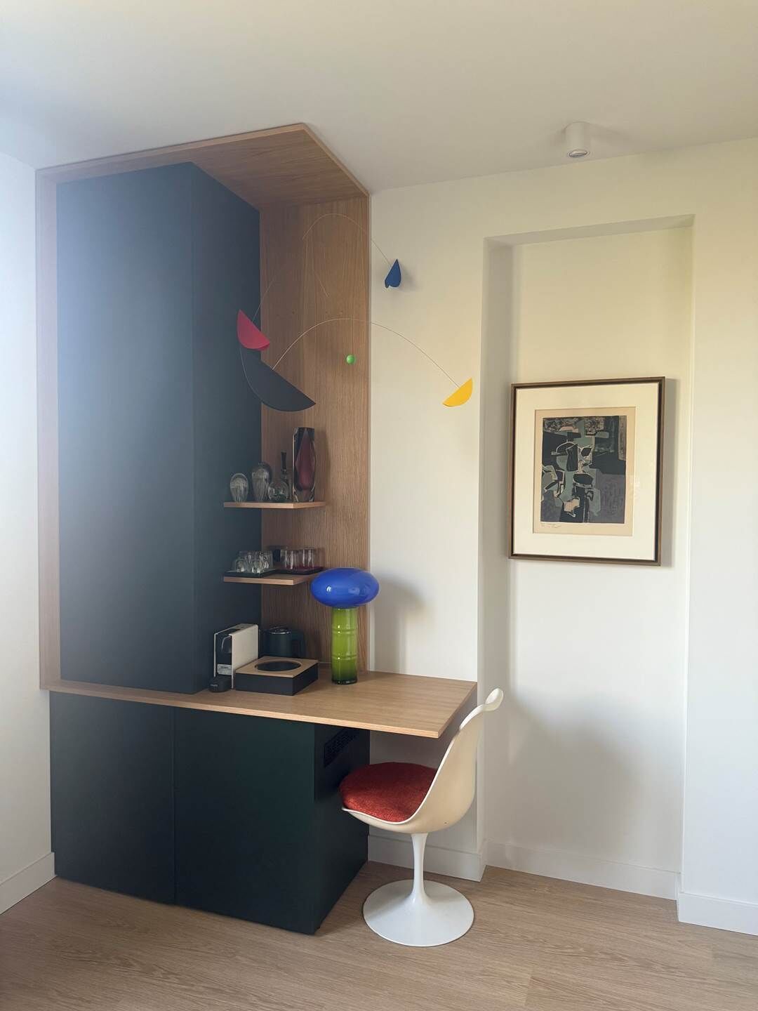 Ein modernes Zimmer mit integriertem Schreibtisch, einem Sessel, einem gerahmten Bild an der Wand und einem Regal mit verschiedenen Gegenständen.