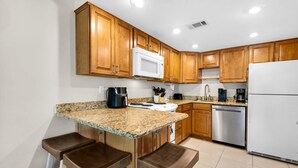 A kitchen with wooden cabinets, a granite countertop, a white microwave, a stainless steel dishwasher, and two bar stools. - Seahorse Siesta (Pensacola Beach)