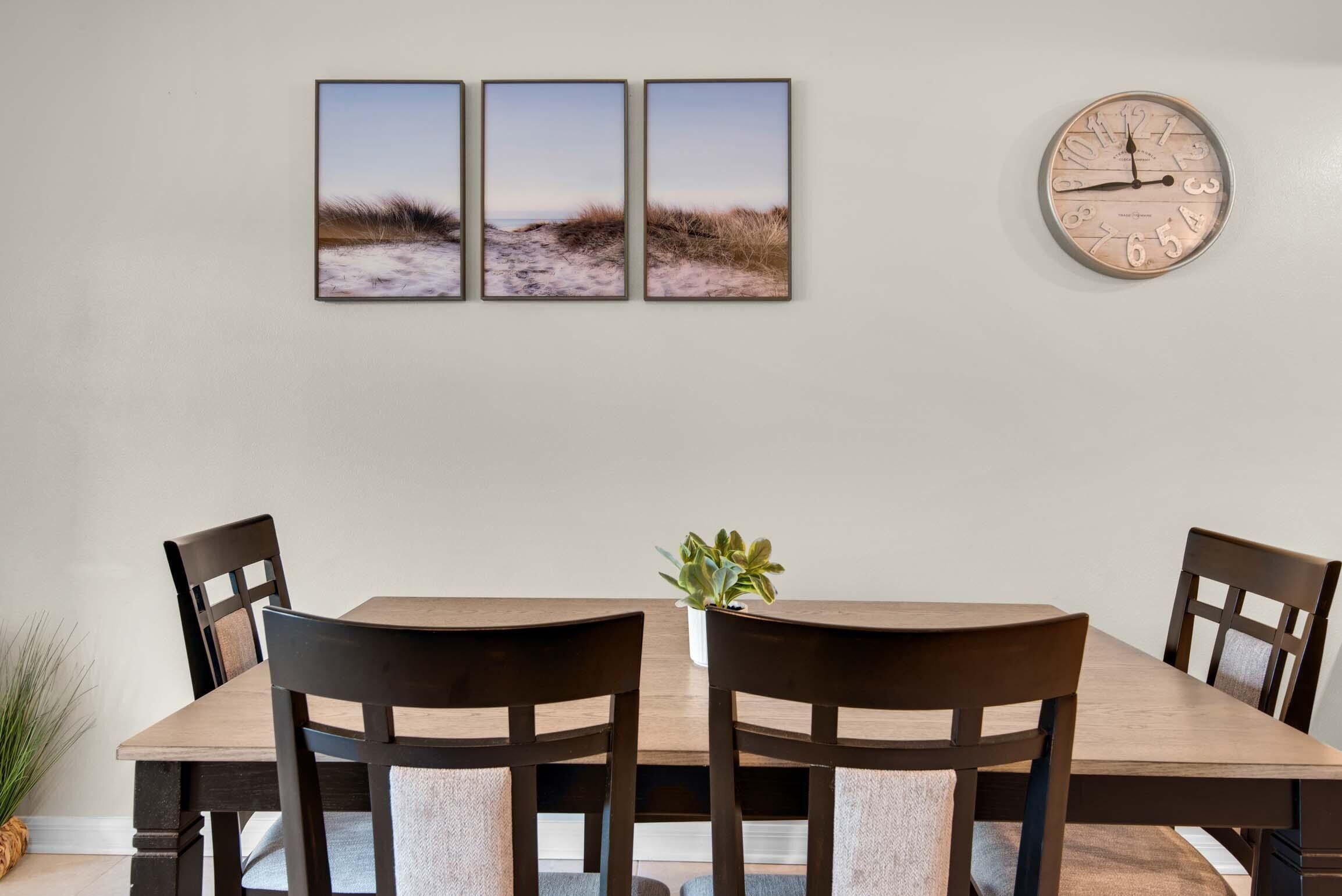 A dining area with a wooden table, four chairs, a clock, and three framed pictures on the wall.