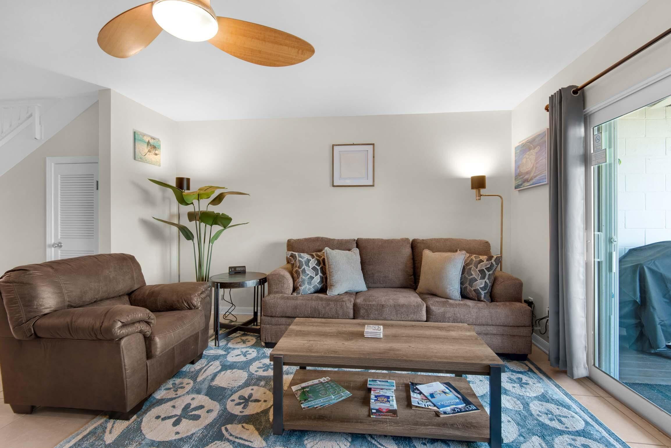 Uma sala de estar com um sofá de couro marrom, uma poltrona de couro marrom, uma mesa de centro de madeira com revistas, um tapete estampado azul e um ventilador de teto.