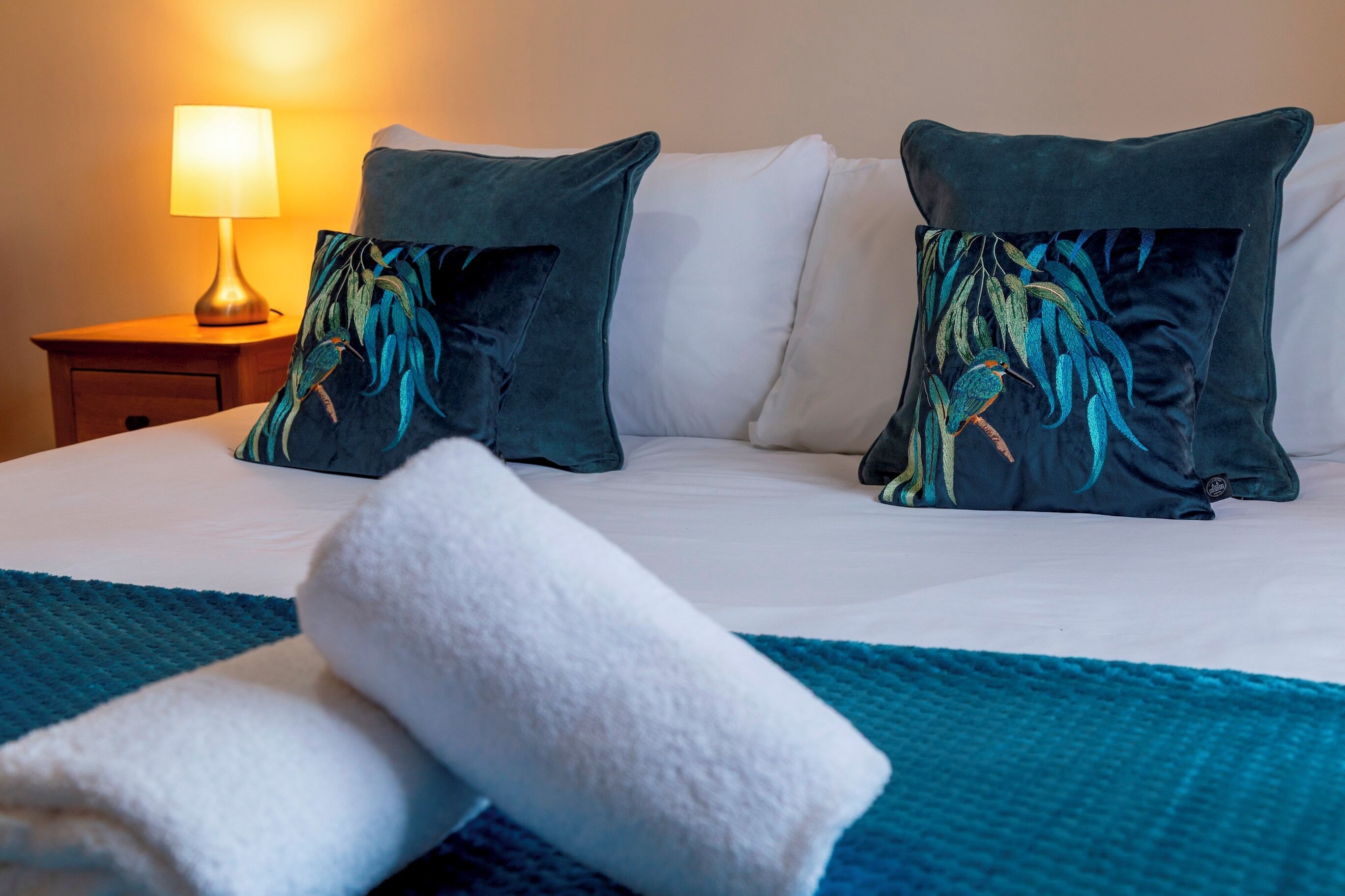 Een net opgemaakt bed met twee decoratieve kussens, een bedlampje en een blauwe gebreide deken.