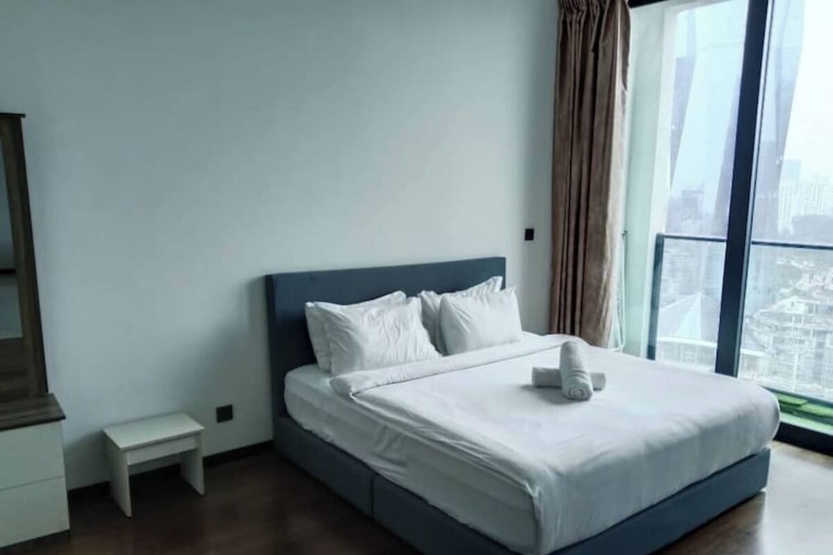 A modern hotel room with a large bed, a small bedside table, and a view of a cityscape through a large window.