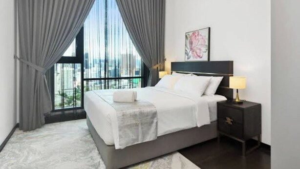 Un dormitorio moderno con una cama grande, una mesita de noche, una lámpara y vistas de la ciudad a través de ventanales del suelo al techo.