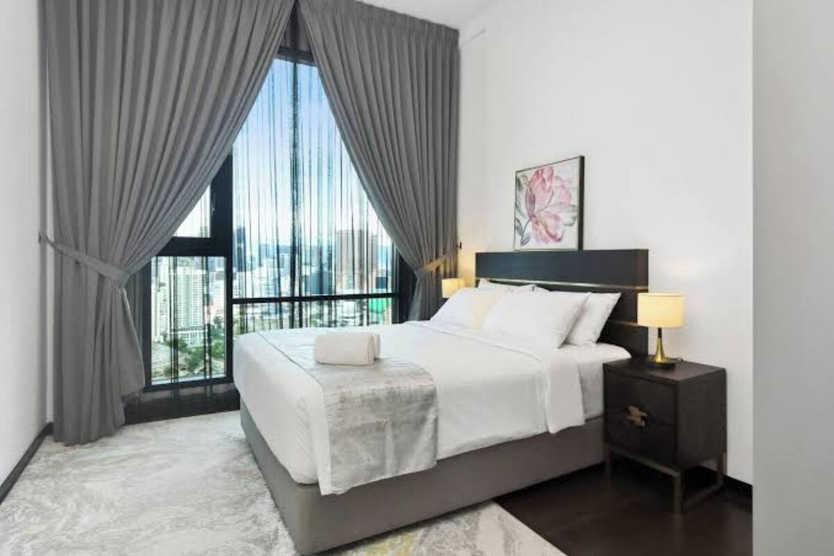 A modern bedroom with a large bed, a nightstand, a lamp, and a view of the city through floor-to-ceiling windows.