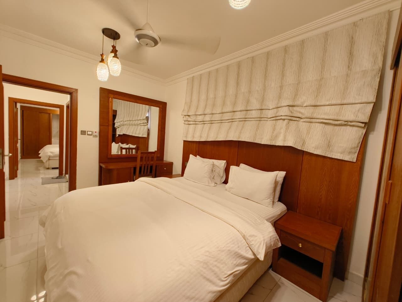 A bedroom with a large bed, wooden headboard, a bedside table, a nightstand, a mirror, and a ceiling light fixture.