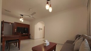 Uma sala de estar com sofá, mesa de centro de madeira, área de refeições com televisão e ventoinha de teto.