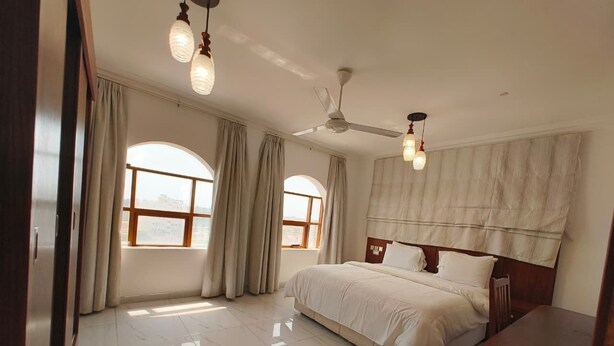 Um quarto espaçoso com uma cama grande, duas janelas com cortinas, um ventilador de teto e uma mesa de cabeceira de madeira.