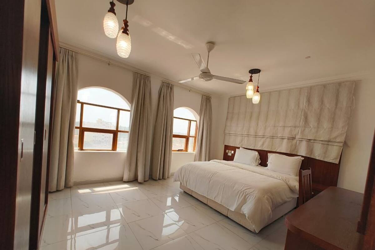 Un dormitorio espacioso con una cama grande, dos ventanas con cortinas, un ventilador de techo y una mesita de noche de madera.