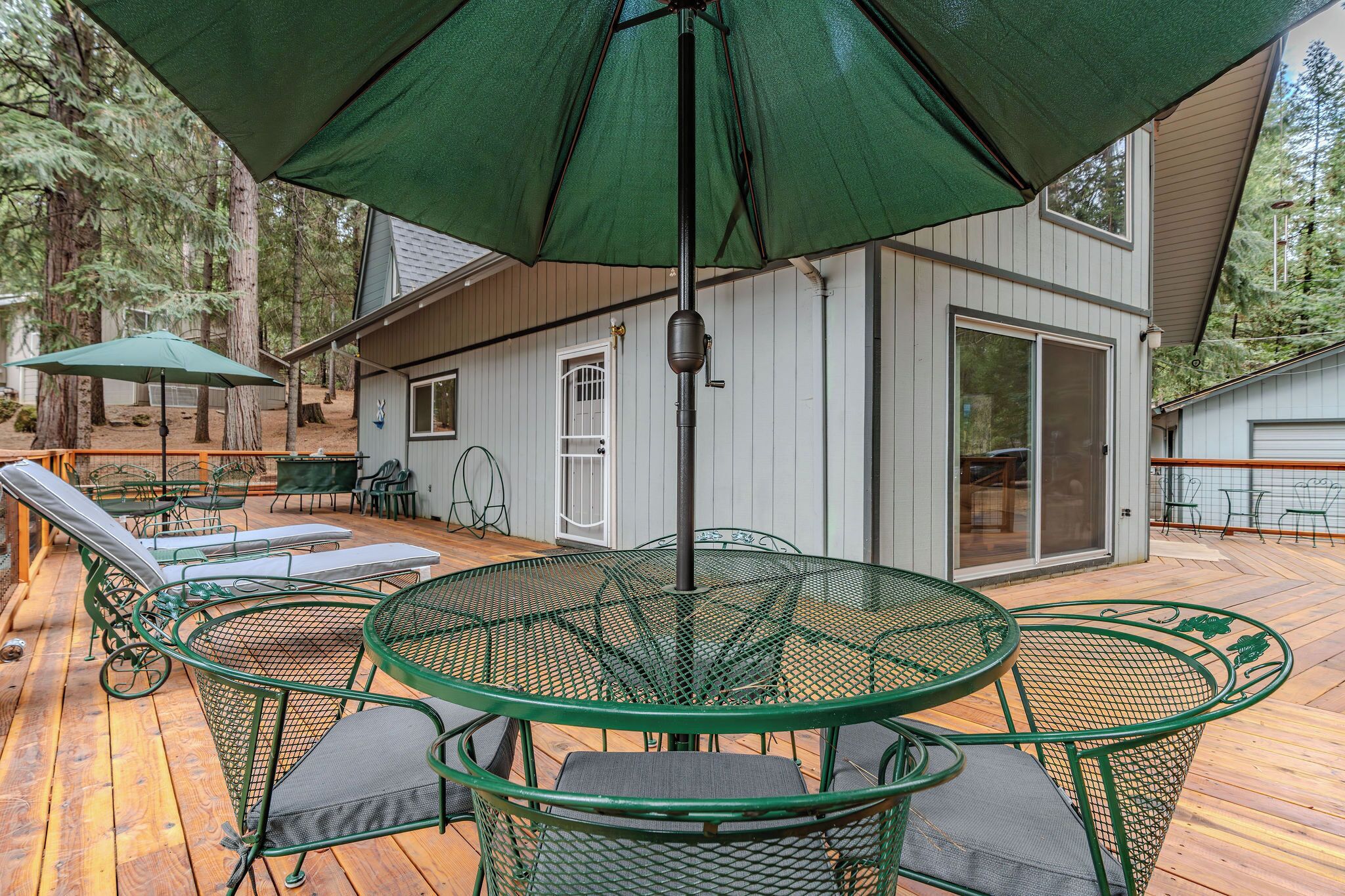 Una terraza de madera con mesas y sillas de metal verde, un paraguas grande y una casa con puerta corrediza de vidrio.