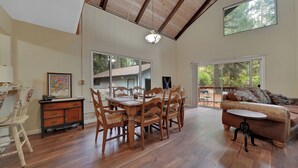 A spacious living area with a dining table, wooden chairs, a sofa, and large windows offering views of the outdoors.
