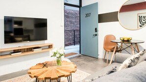 A modern living room with a flat-screen TV, a round wooden coffee table, a small side table with a vase, a blue door with the number 207, a round table with a bowl, and a large mirror on the wall. - The Scarlet – Midtown Apt w/ Balcony & Skyline (Atlanta)