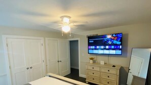 Een slaapkamer met een bed, een dressoir, een spiegel, een televisie en een plafondventilator.
