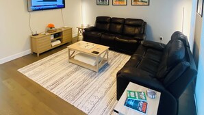 Een woonkamer met een zwarte leren bank, een houten salontafel, een aan de muur bevestigde televisie en een bijzettafeltje met een lamp.