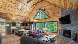 Ein geräumiges Blockhütten-Interieur mit Holzbalkendecke, Kamin aus Naturstein, einem großen Fernseher darüber und einer gemütlichen Sitzecke mit Sofa und Couchtisch.