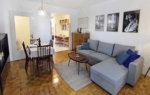 Een woonkamer met een grijze bank, een eettafel met stoelen, een kroonluchter en beelden in lijsten aan de muur.