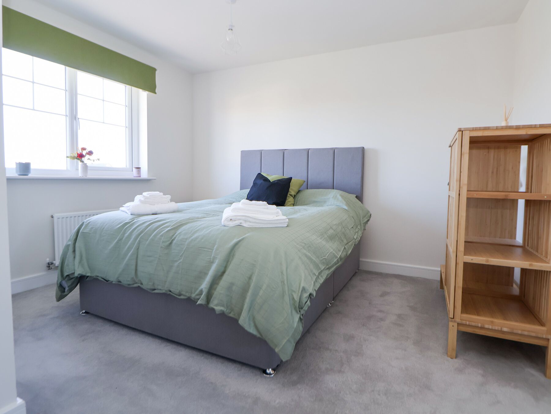 A neatly made bed with a grey headboard, a wooden bookshelf, and a radiator on the wall.