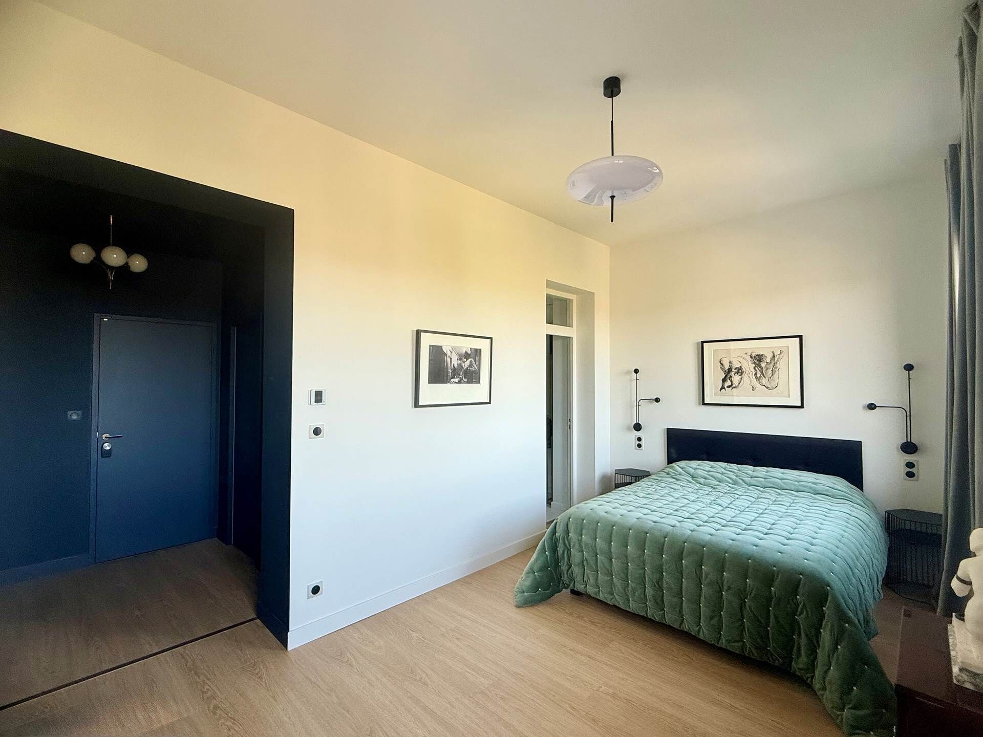 A bedroom with a bed, a door, a light fixture, and two framed pictures on the wall.