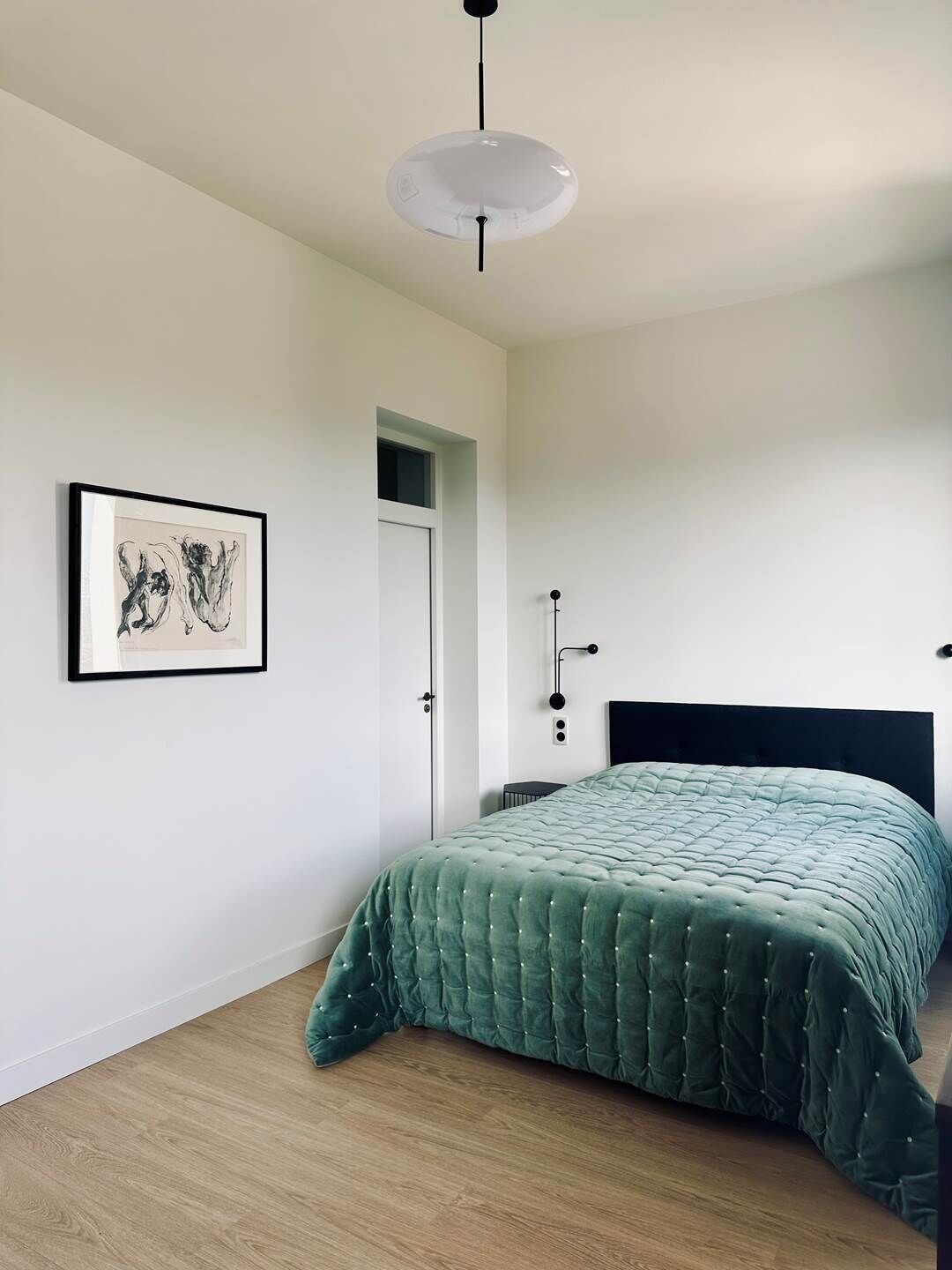 A minimalist bedroom with a green bedspread, a framed abstract artwork, a white door, and a pendant light.