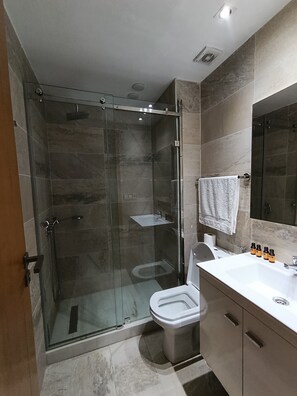 A modern bathroom with a glass-enclosed shower, a toilet, and a sink with toiletries. - La Résidence La Rambla (Fnideq)