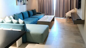 Ein modernes Wohnzimmer mit einem Ecksofa, einem Couchtisch, einem an der Wand montierten Fernseher und einem großen Fenster mit Vorhängen.