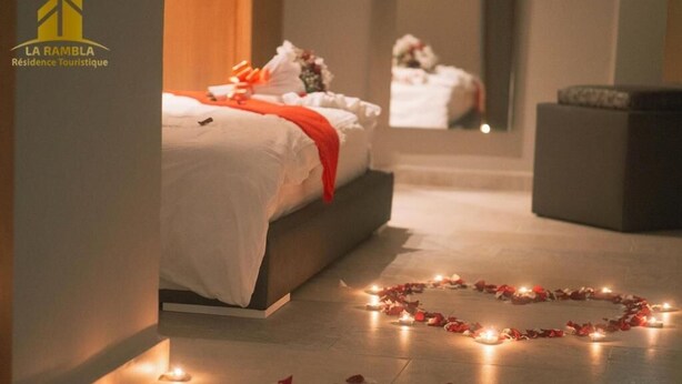 Ein Hotelzimmer mit einem Bett, einer herzförmigen Anordnung aus Kerzen und Rosenblättern auf dem Boden sowie einem Spiegel, der den Raum reflektiert.