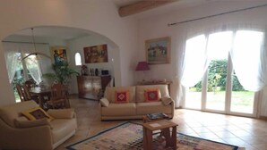 A living room with a sofa, armchair, coffee table, and a patterned rug. There is a dining area with chairs and a table, and a painting on the wall.