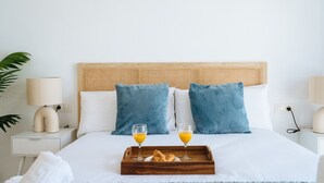Een netjes opgemaakt bed met een houten hoofdbord, twee blauwe kussens en een dienblad met croissants en glazen sinaasappelsap.