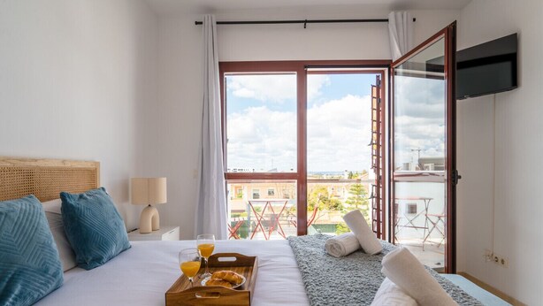 Hotelkamer met een bed, nachtkastje met een lamp, dienblad met croissants en sinaasappelsap, raam met uitzicht op gebouwen en een ladder buiten, en een aan de muur bevestigde televisie.