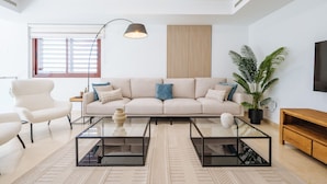 Een moderne woonkamer met een beige bank, twee fauteuils, een glazen salontafel, een houten tv-meubel en een grote potplant.