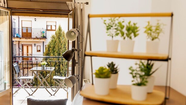 Un balcón con una mesa y sillas, una estantería con plantas en macetas y vistas a un edificio con balcones.