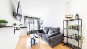 Una sala de estar moderna con un sofá gris, una mesa de centro y un televisor montado en la pared. Hay plantas en estanterías y una ventana con cortinas.