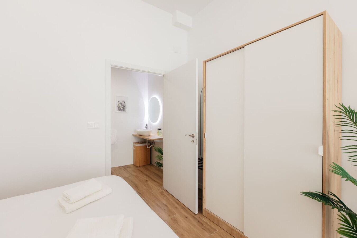 Un dormitorio moderno con piso de madera, una cama con sábanas blancas, un armario blanco y un baño con tocador y espejo de madera.