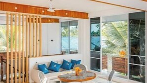 Ein modernes Wohnzimmer mit Holzbalkendecke, einem weißen Sofa mit blauen Kissen, einem Glastisch und Blick ins Freie durch große Fenster.