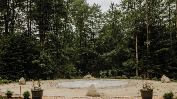 A circular outdoor area with a sandy ground, surrounded by trees. There are several potted plants and rocks placed around the perimeter.
