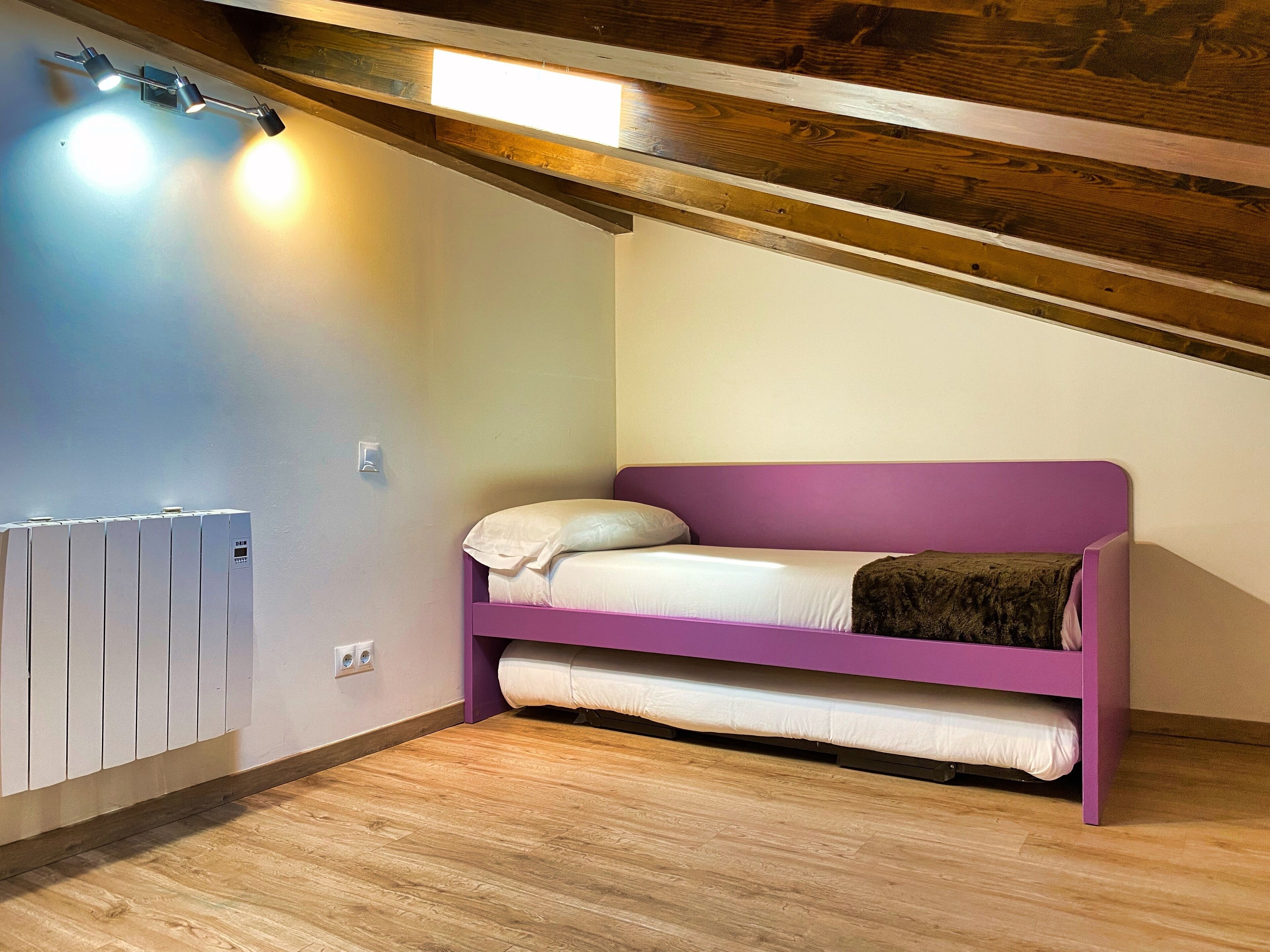 A small, single-bed room with a purple headboard, a white mattress, and a brown blanket. There is a white radiator on the left and a wooden ceiling with exposed beams.