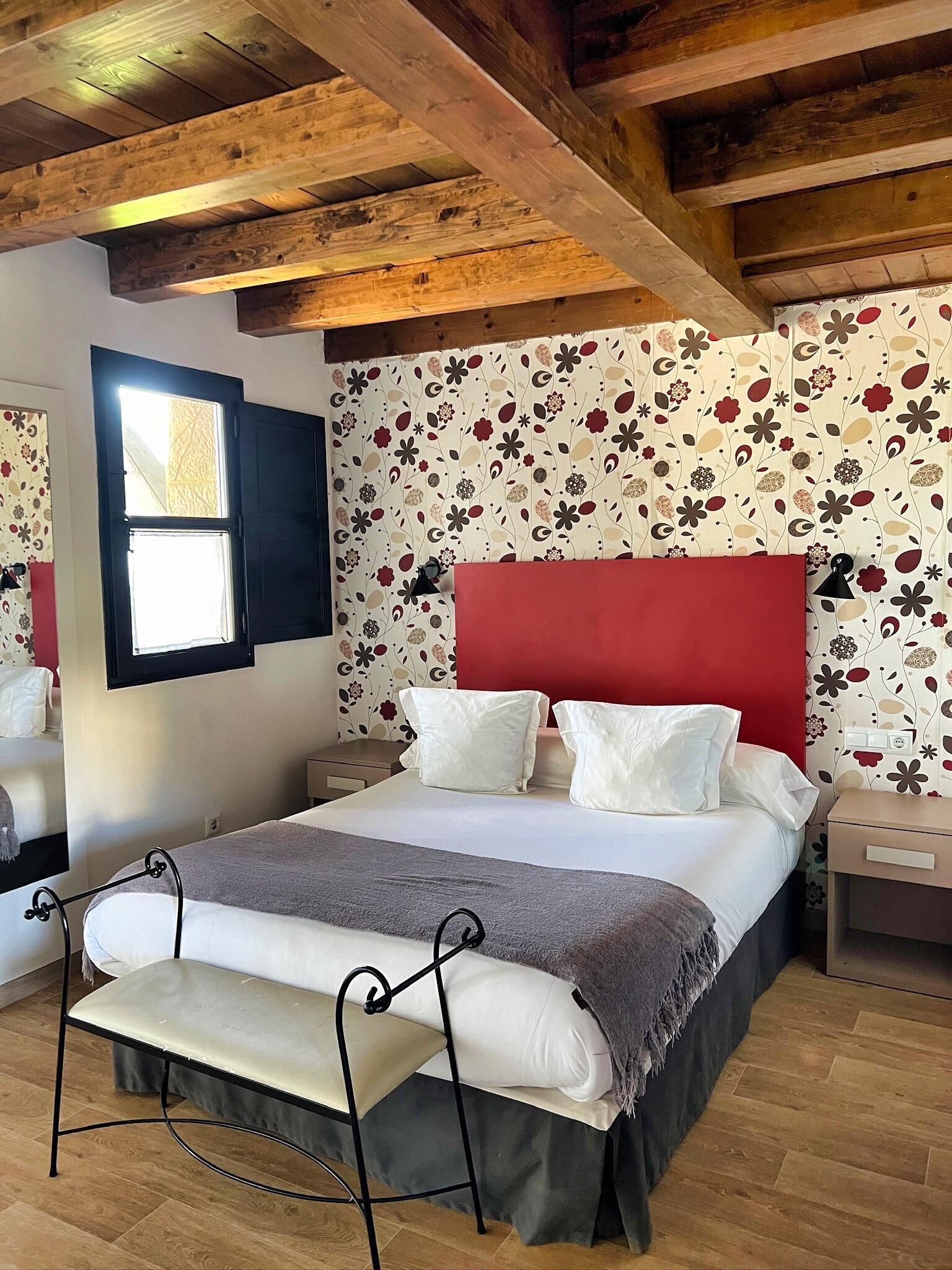 A bedroom with a wooden ceiling, a bed with white bedding and a red headboard, a bedside table, and a window with a view.