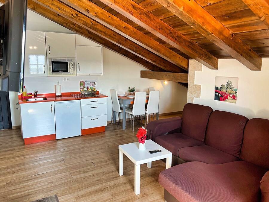 Una cocina moderna con armarios blancos y encimera roja, una zona de comedor con sillas blancas, y una sala de estar con sofá marrón y una mesita blanca.