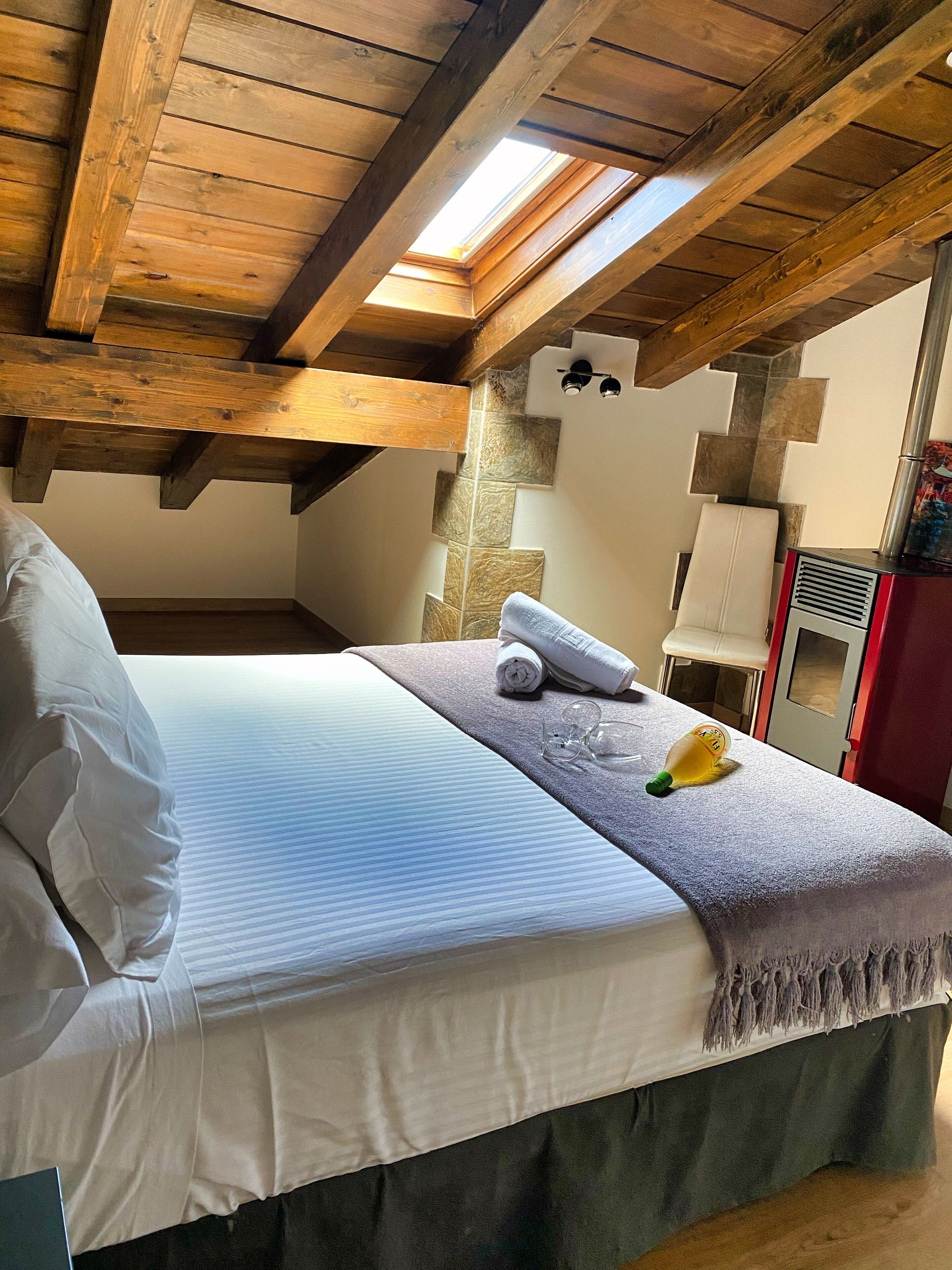 A bedroom with a sloped ceiling, wooden beams, a bed with white linens, a towel, and a bottle of lotion.