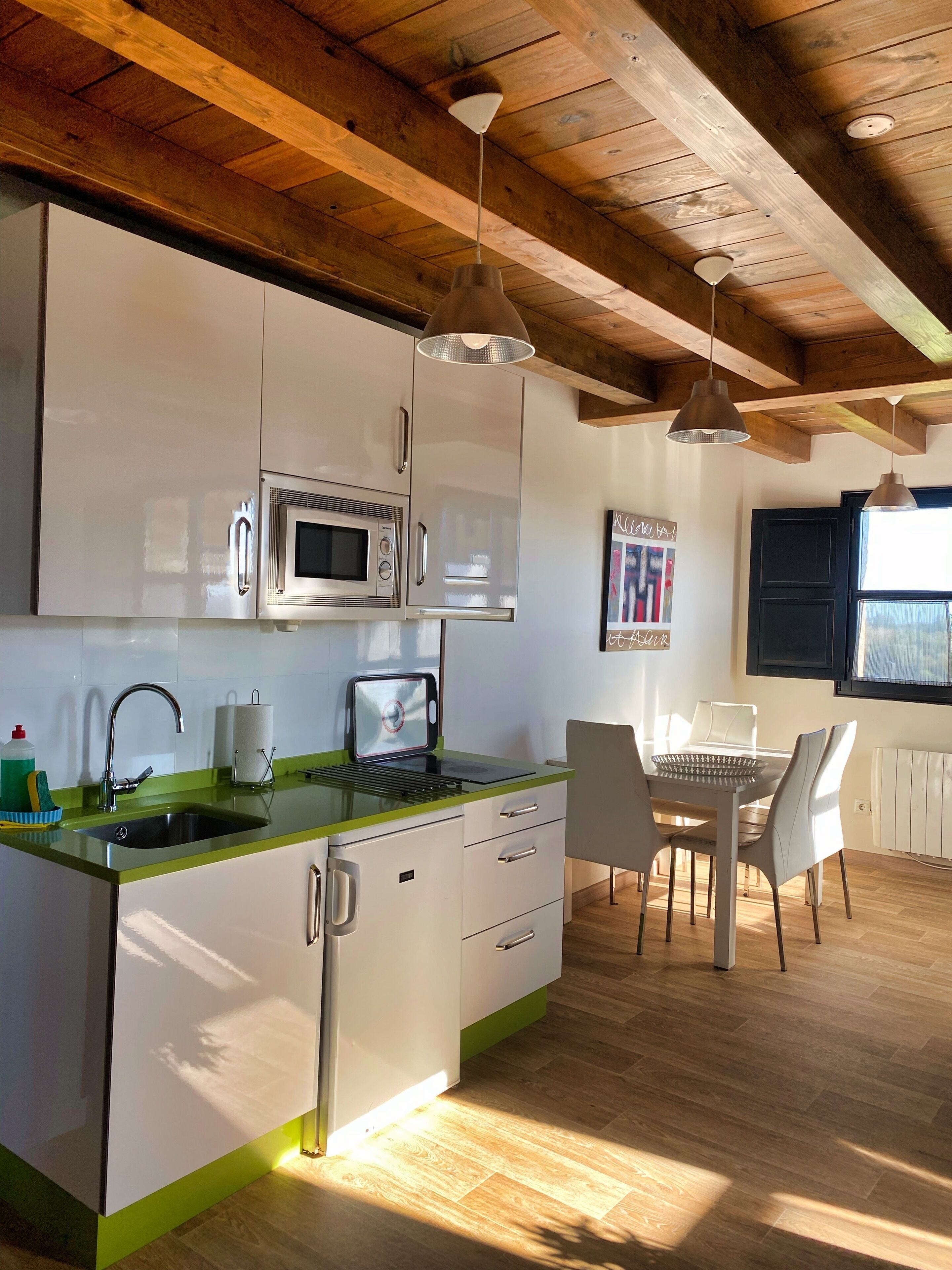Una cocina moderna con armarios blancos, encimera verde y microondas integrado. Hay una zona de comedor con mesa y sillas, y un ventanal que proporciona luz natural.