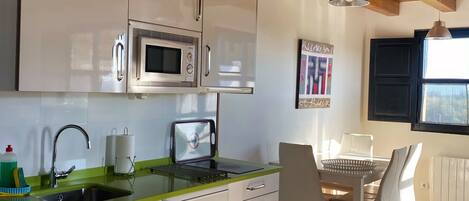 Una cocina moderna con armarios blancos, encimera verde y microondas integrado. Hay una zona de comedor con mesa y sillas, y un ventanal que proporciona luz natural.