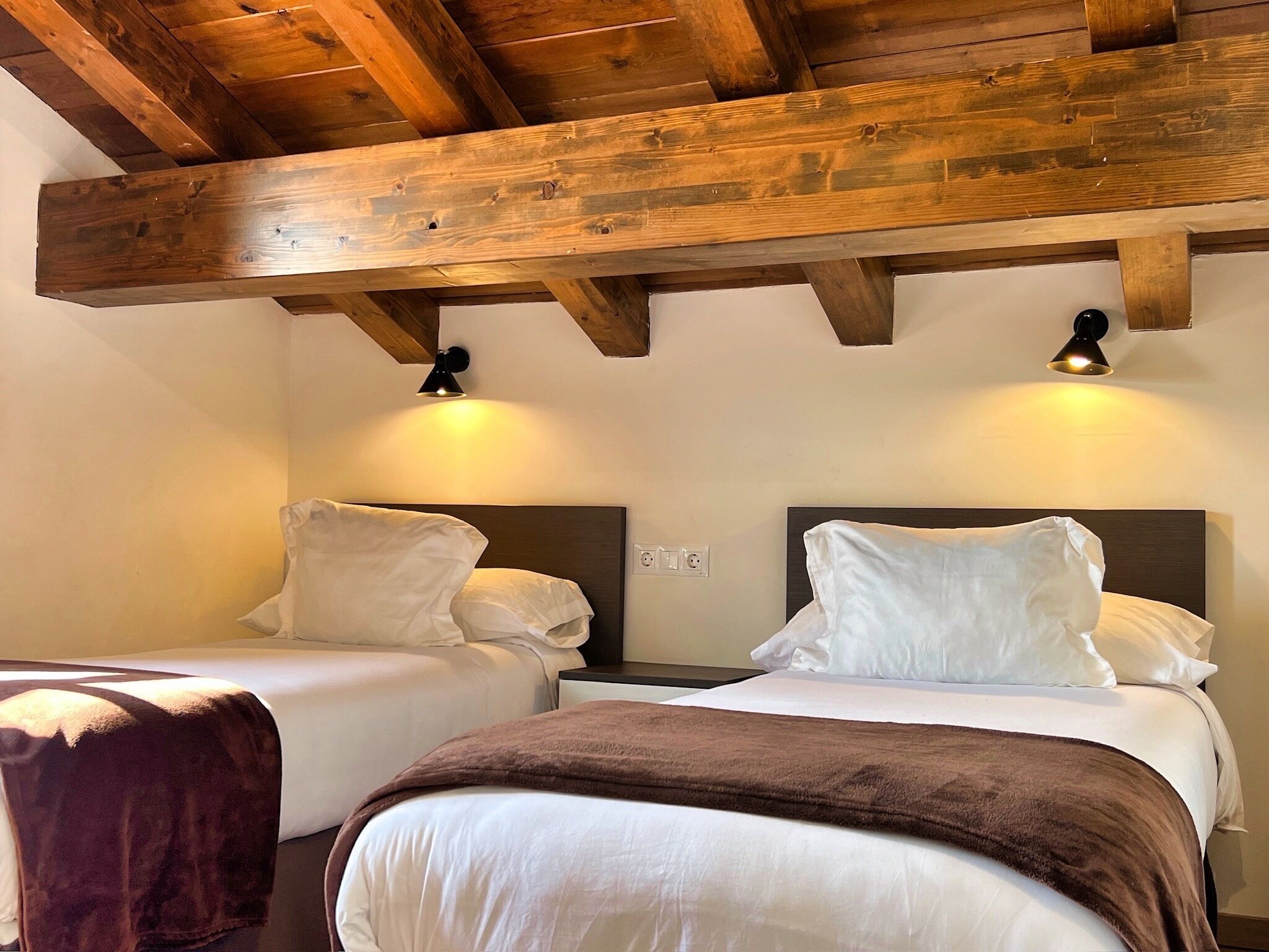 Ett hotellrum med två sängar, synliga träbjälkar i taket och väggmonterade lampor.