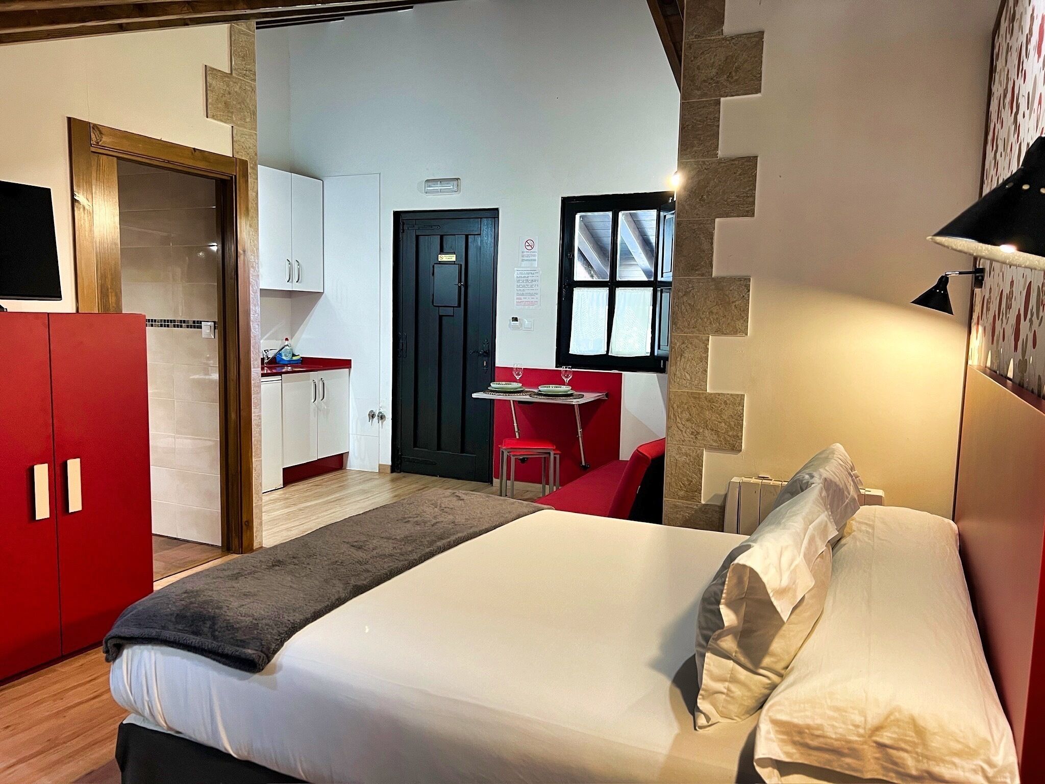 A bedroom with a bed, a red wardrobe, a small kitchenette, and a dining area.