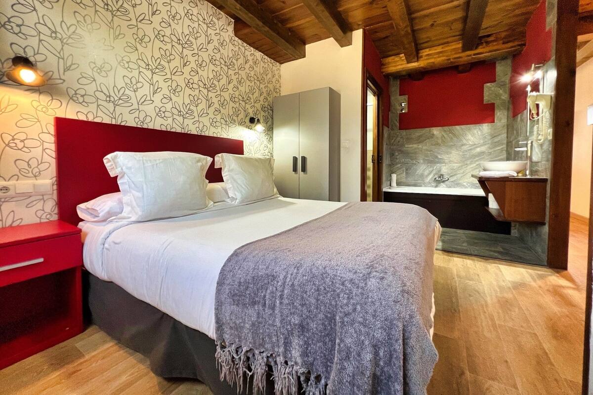 Una habitación de hotel con cama, una mesita de noche roja, un armario, un baño con lavabo y espejo, y techo de madera.