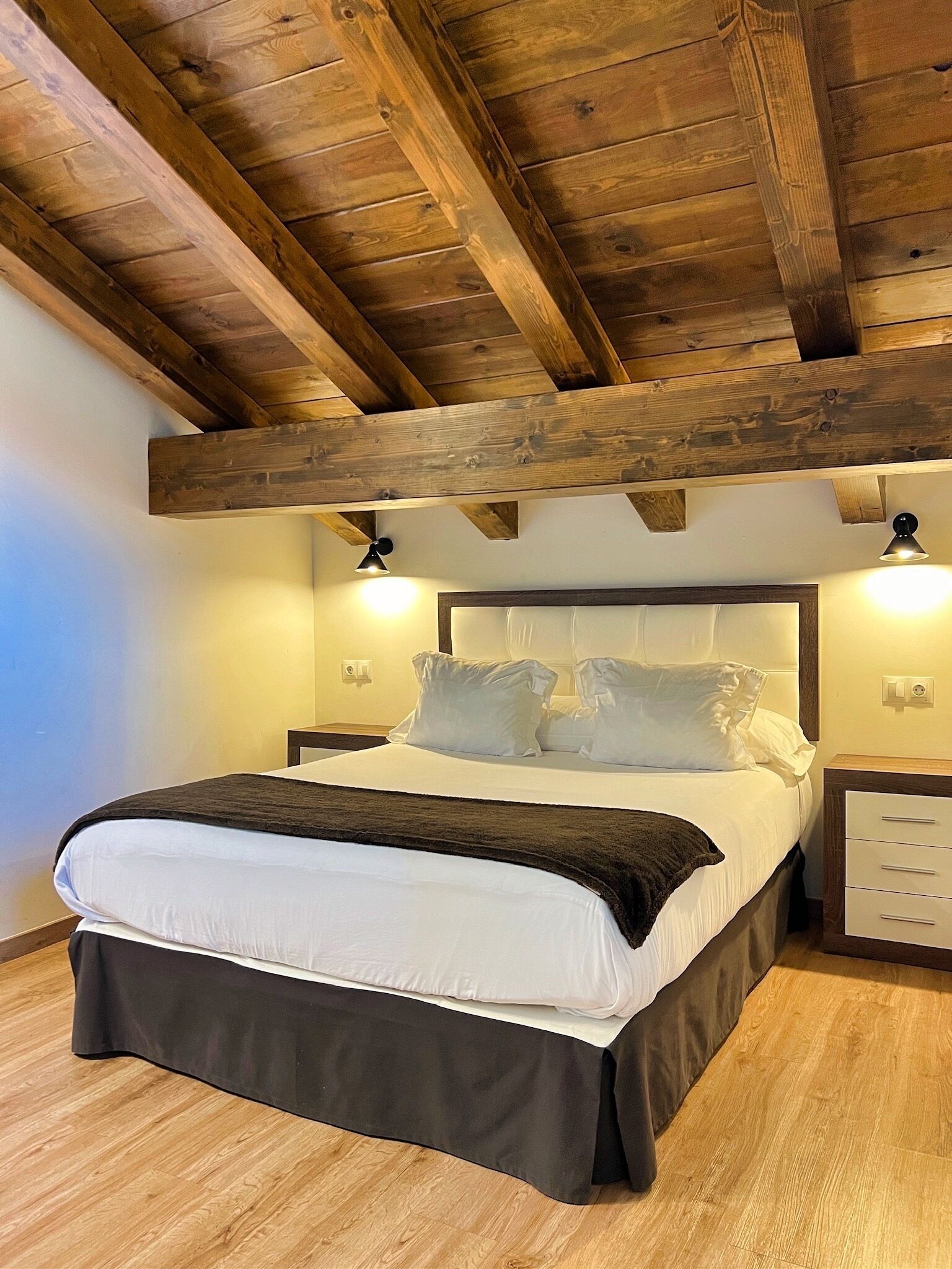 Een slaapkamer met een houten plafond, een bed met wit beddengoed, twee nachtkastjes en een nachtkastje met lades.
