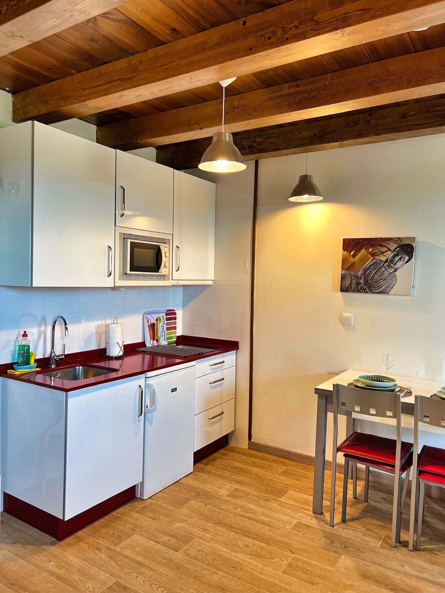 A compact kitchen with white cabinets, a built-in microwave, and a red countertop. There is a dining area with a table and chairs, and a hanging light fixture above.
